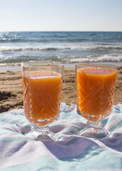 Two glasses with fresh orange juice on a beach. Golden sand and blue sea waves on background. Summer day morning. 