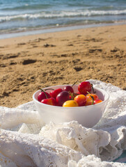 White bowl with sweet cherries on a beach towel. Sunny day on the beach. Blue calm sea waves, golden sand, idyllic summer scenery. 