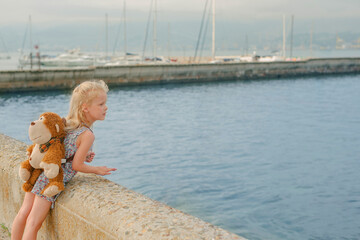 Fototapeta na wymiar little girl with long white hair in a dress with a monkey backpack looks into the distance on the pier of the sea