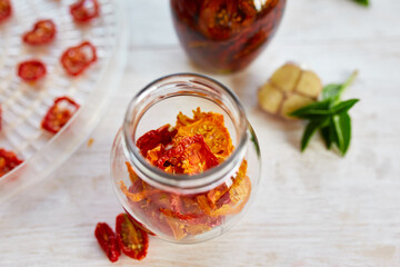 Homemade sun dried tomatoes with herbs, garlic in olive oil in a glass jar on white wooden background. Top view. Print for kitchen