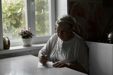 An elderly woman writes at home on a piece of paper. Hobbies of pensioners. Remote communication, sending emails. The process of writing a note