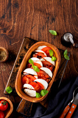 Caprese salad Italian food with ripe tomatoes, fresh garden basil and mozzarella cheese in olive wooden bowl on old rustic background. Top view flat lay