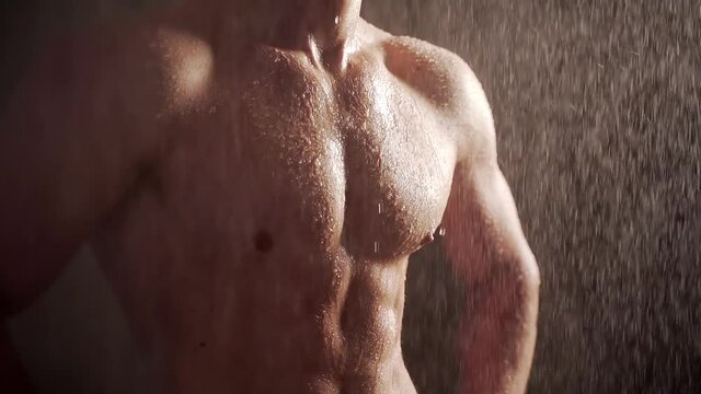 Close up body young man muscular guy bodybuilder on which water pours Portrait of Sexy Male Falling rain or shower on a dark background. Concept: sport, strong healthy body and motivation