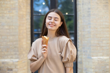 close-up of a smiling brunette girl with closed eyes eating ice cream in a waffle cup against a brick wall background