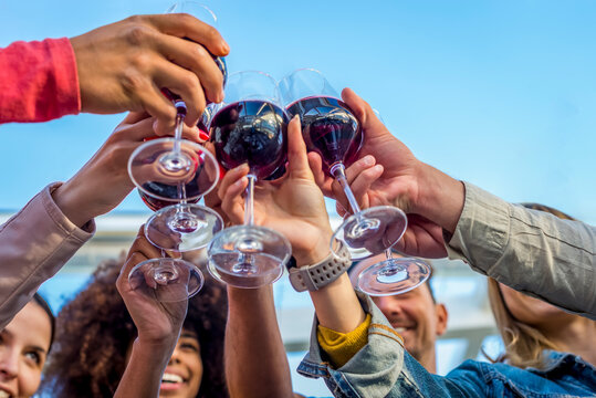 Low Angle Cropped View Of A Diverse Group Of Friends In A Party Making A Celebratory Toast Clinking Wine Glasses. Happy People Having Fun At Summer Sunset Happy Hour. Focus On Glasses From Below.