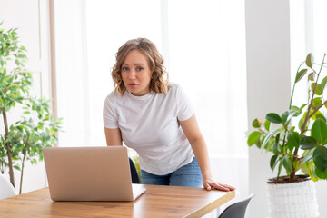 Business woman using laptop standing near desk white office interior with houseplant looking at camera