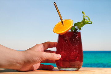 Hand holding rum cocktail with cranberry juice on the tropical beach. Glass of alcoholic refreshing drink decorated orange slice and mint. Beach bar, summer sea resort concept.