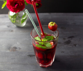 Cocktail with red wine, ice, strawberry, mint in high glass with straw on black background and rose flowers bouquet in vase