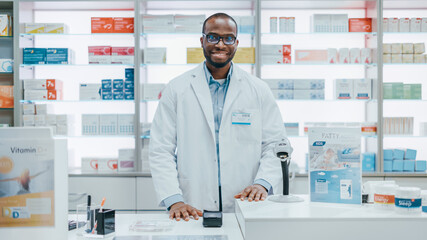Naklejka na ściany i meble Pharmacy Drugstore Checkout Counter: Portrait of Handsome Professional Black Male Pharmacist Wearing White Lab Coat, Looks at Camera, Smiles. Shelves with Medicine Packages, Health Care Products