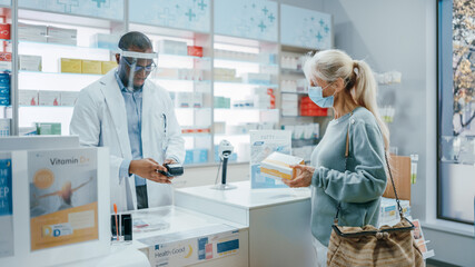 Obraz premium Pharmacy Drugstore Checkout Counter: Professional Black Pharmacist Wearing Face Shield Sells Medicine to Senior Female Customer, who is wearing Face Mask, Use Contactless Payment. Coronavirus Safety
