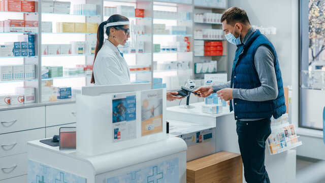 Pharmacy Drugstore Checkout Counter: Female Pharmacist Wearing Face Shields Selling Medicine Package, Customer with Face Mask Using NFC Smartphone with Contactless Payment Terminal and Credit Card