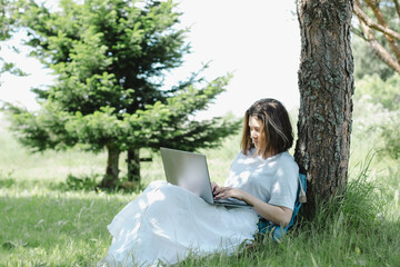 Nice teenage girl siting near tree in summer park with open laptop in her hands and studying. Concept of distance learning