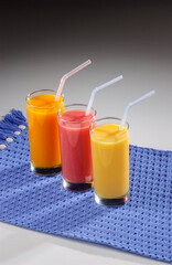 Three glasses of fruit juice with plastic straws over blue placemat on gray gradient background.