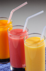 Three glasses of fruit juice with plastic straws over blue placemat on gray gradient background.