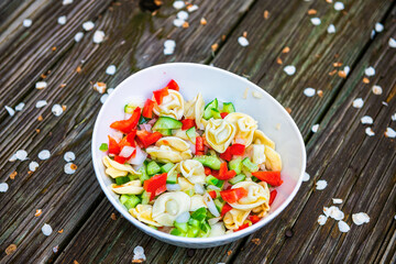 Tortellini pasta salad on table wooden deck with cherry blossom fallen petals on floor and cucumber bell pepper slices in bowl