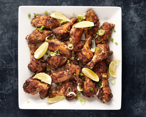 Peppercorn Chicken Wings with Scattered Herbs and Lemon Wedges on a White Square Plate on a Dark Grey Background
