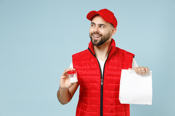 Delivery guy employee man in red cap white T-shirt vest uniform work as dealer courier tablets aspirin pills in bottle, blank paper bag isolated on pastel blue color background studio Service concept - obrazy, fototapety, plakaty