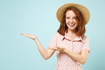 Happy young redhead woman 20s wear casual pink dress straw hat pointing finger hand aside on workspace copy space mockup promo commercial area isolated on pastel blue color background studio portrait