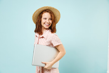 Side view smiling happy young redhead curly woman 20s wears casual pink dress straw hat hold laptop pc computer under her hand look saide back isolated on pastel blue color background studio portrait.