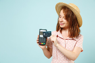 Young redhead woman 20s wears casual pink dress straw hat hold wireless modern bank payment terminal to process acquire credit card payments isolated on pastel blue color background studio portrait