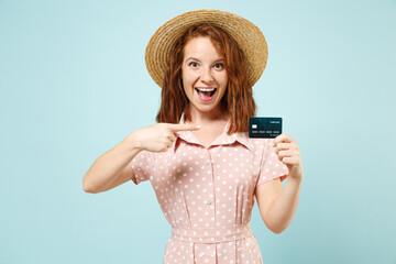 Smiling excited happy young redhead curly woman 20s hold in hand credit bank card pointing on it wears casual pink dress straw hat look camera isolated on pastel blue color background studio portrait