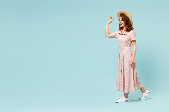 Full size body length side view young redhead curly woman 20s wear casual pink dress straw hat walk strolling meet greet waving hand as notices someone isolated on pastel blue color background studio.