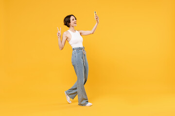 Fototapeta na wymiar Full length young happy friendly woman 20s with bob haircut in white tank top shirt doing selfie shot on mobile phone post photo on social network show victory gesture isolated on yellow background.