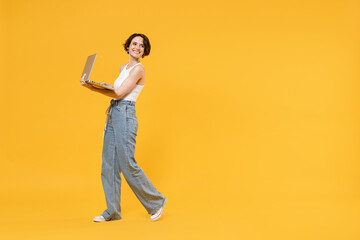 Fototapeta na wymiar Full length side view young smiling freelancer fun woman 20s wear white tank top shirt using laptop pc computer chat online browsing internet walk isolated on yellow color background studio portrait.