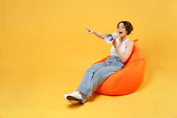 Young smiling fun woman 20s in white tank top shirt scream shout in megaphone point index finger aside on worksapce area sit in orange bag chair isolated on yellow color background studio portrait