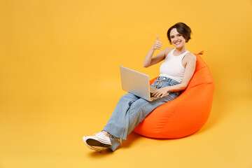 Young fun woman 20s with bob haircut in white tank top shirt hold laptop pc computer chat online browsing surfing internet sit in orange bag chair show thumb up gesture isolated on yellow background