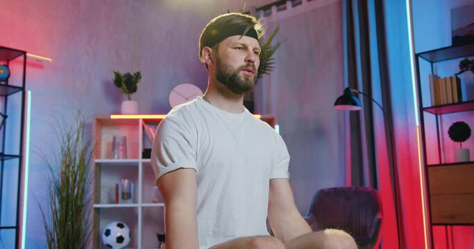 Attractive strong active young bearded man in sportswear sitting doing stretching exercises with resistance band at home in the evening