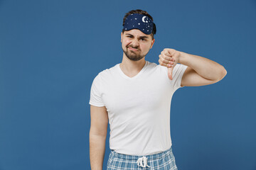 Naklejka premium Young sad displeased dissatisfied man 20s in pajamas jam sleep mask white t-shirt rest relax at home show thumb down dislike gesture isolated on dark blue background Bad mood night bedtime concept