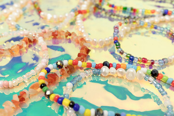 Closeup of necklaces and bracelets made from colorful beads and pearls on a turquoise holographic background.