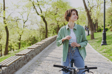 Young woman 20s in green jacket jeans riding bicycle bike in city spring park outdoors, look aside drink clear fresh pure water from transparent glass of water People active healthy lifestyle concept - Powered by Adobe