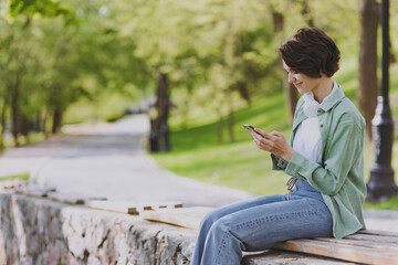 Young smiling happy woman 20s in casual green jacket jeans sit on bench in city spring park outdoors using hold mobile cell phone chat online in social network People active urban youthful concept
