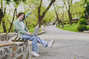 Full size young fun happy student freelancer woman in green jacket jeans sit on bench in spring park outdoors rest use laptop pc computer do winner gesture clench fist People urban lifestyle concept.