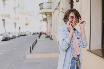Fototapeta premium Young smiling caucasian happy woman 20s wearing casual jeans clothes eyeglasses talk speby mobile cell phone strolling walk go down city street outdoors People youth active urban lifestyle concept