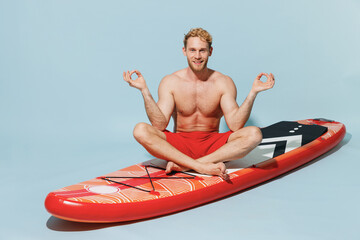 Full length young blond sporty man in red shorts swimsuit sit on sup board spread hands in yoga om gesture relax meditate isolated on pastel blue background Summer vacation sea rest sun tan concept.