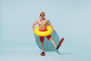 Full length young sporty man in red shorts swimsuit diving mask relax near pool hold inflatable ring flippers raise up leg isolated on pastel blue background. Summer vacation sea rest sun tan concept