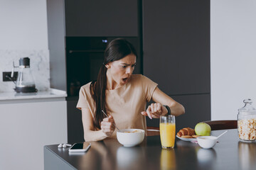 Young disappointed housewife woman 20s in t-shirt eating late breakfast in morning look at smart...
