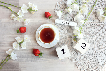 Calendar for July 12 : cubes with the number 12, the name of the month of July in English, a cup of tea on a gray openwork napkin, scattered flowers of bluebells and strawberries, top view