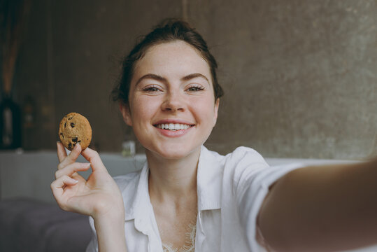 Close up young smiling happy woman in white clothes glasses sit on grey sofa indoors apartment do selfie shot on mobile phone hold eating cookie Rest on weekend leisure quarantine stay home concept.