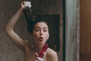 Half naked topless young cool woman 20s take shower hold bottle of shampoo gel in bathroom show wet hair doing morning routine in brown background. Skin care healthcare cosmetic procedures concept
