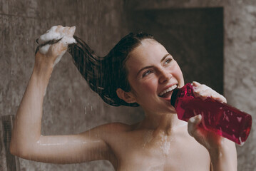Half naked topless young woman 20s taking shower sing song in microphone bottle of shampoo hold wet...