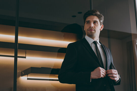 Bottom view young successful employee agent business man corporate lawyer 20s wear formal black suit shirt tie look aside illuminated indoors apartment dark wall. Achievement career lifestyle concept.