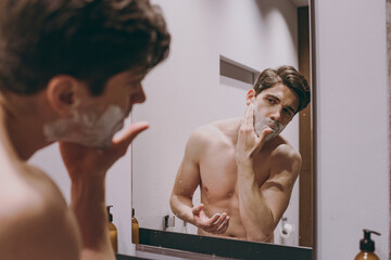 Young handsome half naked topless caucasian brunette man 20s reflected in mirror shaving beard...
