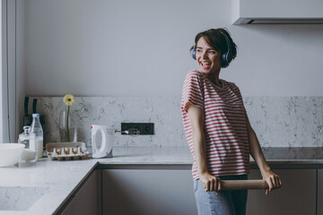 Young excited housewife woman 20s wear casual clothes striped t-shirt headphones holding rolling pin listen to music cooking food in light kitchen at home alone. Healthy diet bakery lifestyle concept.