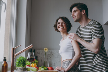 Young side bottom view minded smiling happy couple two married woman man in casual t-shirt clothes looking aside hug wife cook food in light kitchen at home together Healthy diet lifestyle concept