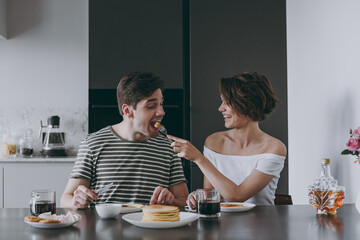 Young cheerful happy couple two woman man in casual t-shirt clothes sit by table eat pancake with maple syrup feed boyfriend cook food in light kitchen at home together Healthy diet lifestyle concept