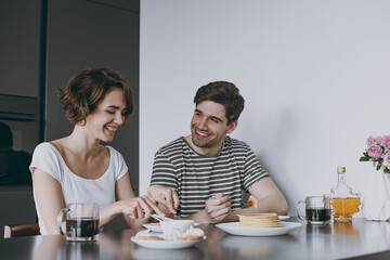 Fototapeta na wymiar Young satisfied happy couple two caucasian woman man in casual t-shirt clothes sit by table eat pancakes with maple syrup cook food in light kitchen at home together Healthy diet lifestyle concept